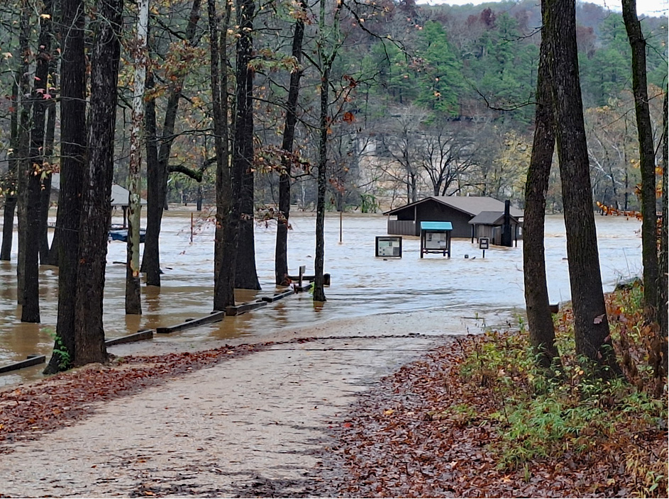 A tree lined dirt road can be seen leading into a flooded campground. Signs and a brown restroom facility can be seen in the center of the flood waters.