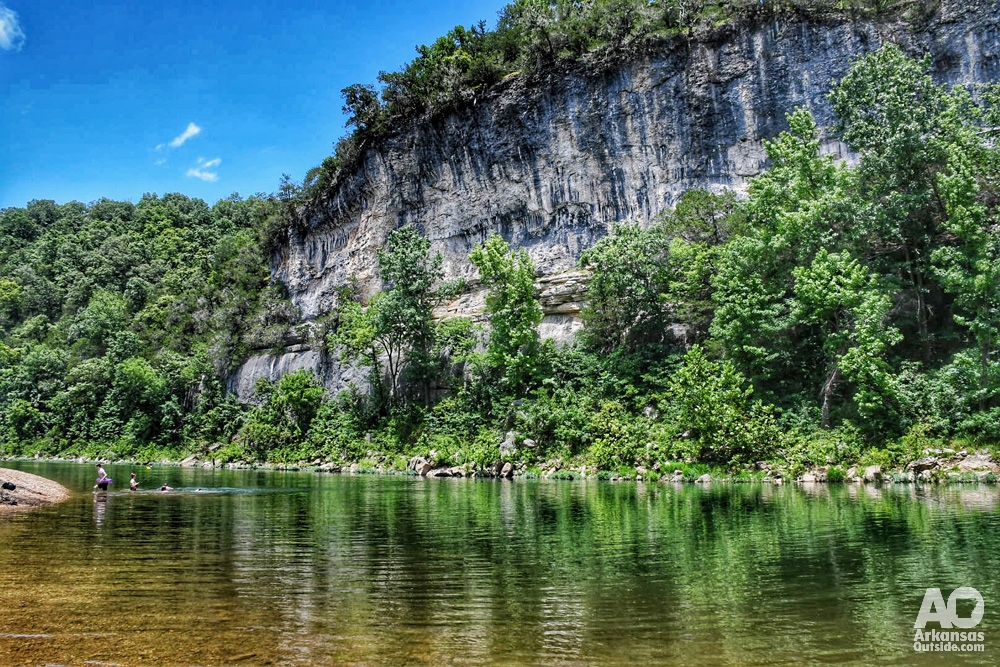 Looking across the Buffalo National River at a tall, rock bluff line while swimmers enjoy the water.