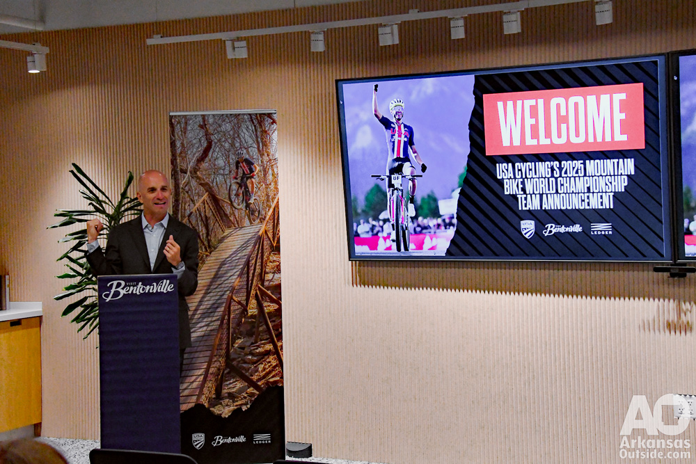 Man speaking at a podium with a video welcome message beside him.
